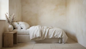 A small bedroom with limewash walls in a warm white and ivory tone, showing the characteristic cloudy, slightly uneven texture of limewash across the full wall surface. Natural morning light shifts the tone from warm ivory in bright spots to soft cream in shadowed areas. Simple linen bedding, natural wood nightstand, a ceramic vase with dried grasses. The room reads as ancient and serene. Editorial interior photography.